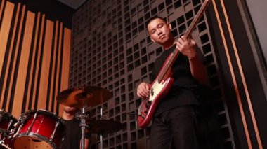 Genç Asyalı bir adam ses geçirmez müzik stüdyosunda bas gitar çalıyor, davulcu kısmen görünür, profesyonel ışıklandırma, odaklanmış atmosfer.