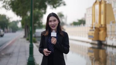 Elinde mikrofon tutan kendine güvenen Asyalı kadın gazeteci, şehirdeki altın anıt ve kanal geçidi dışında haberler sunuyor, resmi takım elbise giyiyor, açık hava çekimi sırasında gülümsüyor.