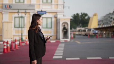 Siyah takım elbiseli genç Asyalı iş kadını caddede yürüyor ve akıllı telefona bakıyor. Gündüz vakti miras mimarisi ve hükümet muhafızlarının yanından geçiyor.