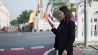 Siyah takım elbiseli Asyalı kendine güvenen bir iş kadını tarihi hükümet binasının önünde yürürken cep telefonuyla konuşuyor ve kol saatini kontrol ediyor.