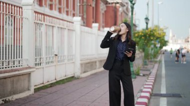 Asyalı iş kadını neşeyle gülümsüyor, kahve içiyor ve kaldırımda akıllı telefon kullanıyor, yoğun sabah rutininde rahatlama ve mutluluk ifade ediyor.