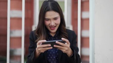 Genç Asyalı iş elbiseli bir kadın metal çitlerin yanında oturuyor, akıllı telefonu yatay olarak tutuyor, odaklanmış ifadesiyle, mobil oyun anından zevk alıyor.