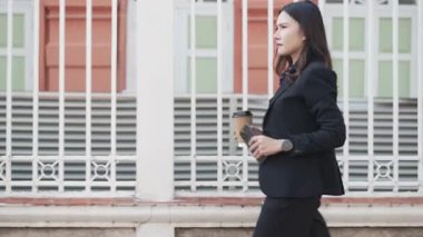 Siyah takım elbiseli Asyalı genç bir kadın kahve fincanı ve akıllı telefonuyla çitin yanında yürüyor, ilerlerken telefona bakıyor, kamera yan görüntüsünü takip ediyor.
