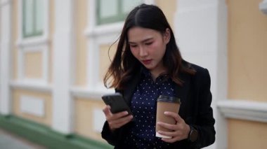 Asyalı iş kadını elinde kahve fincanı ve telefonla konuşuyor ve neşeli yüz ifadesi ve modern yaşam tarzıyla şehirde arkadaşını mutlu bir şekilde karşılıyor.