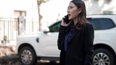 Resmi takım elbiseli genç Asyalı kadın şehir sokağında akıllı telefondan konuşurken gülümsüyor, güpegündüz dostça sohbet ediyor.