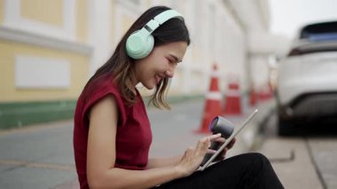 Şehirdeki patikada oturan genç Asyalı kadın, kablosuz kulaklık takıyor, müzik çalarken gülümsüyor ya da tablet üzerinde uygulamalar yapıyor, yeniden kullanılabilir takla atıyor, rahat bir ruh hali içinde.