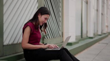 Asyalı kadın binanın dışında yeşil duvarın yanında oturuyor, dizüstü bilgisayar kullanıyor ve kahve içiyor, rahat bir ifadeyle gülümsüyor, şehir merkezinde modern serbest yaşam tarzını temsil ediyor.
