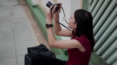 Asyalı mutlu kadın fotoğrafçı kaldırımda oturuyor, kamerayı tutuyor ve gülümsüyor lensi yukarı doğrultuyor, seyahat fotoğrafçılığı sırasında ilham anının tadını çıkarıyor.