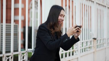 Dışarıda gezici bir oyun oynarken gülümseyen Asyalı genç bir kadın, siyah ceketli, iki eliyle akıllı telefonu tutan, neşe ve konsantrasyonu ifade eden, dijital yaşam tarzını ve şehir gençlik kültürünü temsil eden