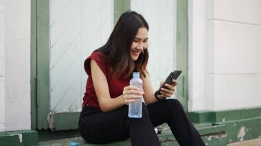 Genç Asyalı kadın dışarıda merdivenlerde oturuyor, gülümsüyor, akıllı telefon kullanıyor ve elinde su şişesi tutuyor, rahat dijital anın tadını çıkarıyor.