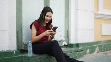 Genç Asyalı bir kadın şehrin dışında oturmuş gülümserken yanında su şişesi olan akıllı telefon kullanıyor sakin ve neşeli bir dijital mola veriyor.