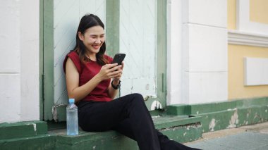 Genç Asyalı kadın akıllı telefon kullanarak gülümsüyor ve şehrin dışında bir adım önde, yanında su şişesiyle yalnız başına gündelik dijital anın tadını çıkarıyor.