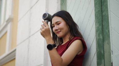 Asyalı kadın dışarıda portatif saç kurutma makinesi kullanıyor, kendine güveni ve rahatlığıyla gülümsüyor, kırmızı bluz ve akıllı saat giyiyor, yaz melteminin ve tazeliğin tadını çıkarıyor.