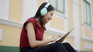 Pastel kulaklık takan genç Asyalı kadın bir şehir binasının yanında otururken tablet kullanıyor, odaklanmış ve gülümsüyor, modern yaşam tarzını ve mobil teknolojisini temsil ediyor..
