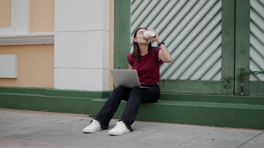 Genç Asyalı kadın dizüstü bilgisayarla ve kahve servisi yaparak gündelik bir şekilde oturuyor. Odaklanma ve rahatlıkla içiyor. Şehir kaldırımında modern yaşam tarzı, özgürlük ve yaratıcılık gösteriyor..