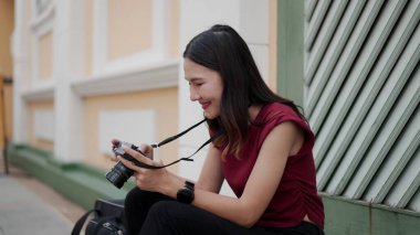 Genç Asyalı bayan gezgin kaldırımda oturuyor, kameradaki fotoğraflara bakıp gülümsüyor, modern şehir seyahatinde gündelik dijital yaşam tarzında yaratıcılık, neşe ve huzur dolu anları gösteriyor..