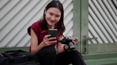 Genç Asyalı bayan gezgin kamerayla dışarıda oturuyor, akıllı telefondan fotoğrafları kontrol ederken gülümsüyor, modern ortamdaki kişisel seyahat hikayesinde neşe, yaratıcılık ve dijital yaşam tarzını gösteriyor..