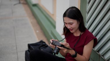 Genç Asyalı bir kadın şehrin kaldırımında tek başına dolaşırken mutlu bir şekilde gülümsüyor ve fotoğraf makinesindeki fotoğrafları kontrol ediyor.