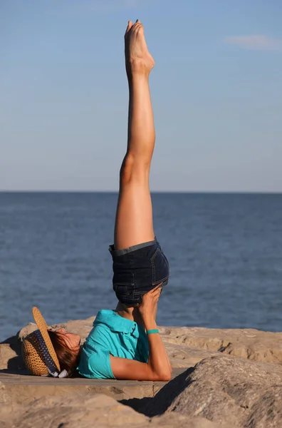 一个身材苗条的小女孩 穿着蓝色T恤 戴着草帽 在海边岩石上做体操锻炼 — 图库照片