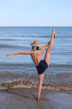 Hasır şapkalı genç atletik kız deniz kıyısında bacağını havaya fırlatıyor.