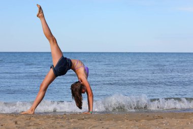 Genç, narin bir kız yazın kafası deniz kıyısında jimnastik yapıyor.
