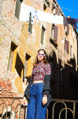 girl with venetian mask wearing jeans trousers posing near houses with clothes hanging out to dry in the sun in Venice in Italy clipart
