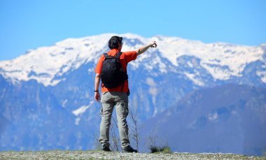 young Man hiker with black backpack standing on a mountain trail looking at snowcapped peaks clipart