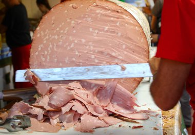 Bıçaklı bir şef, İtalyan dilinde MORTADELLA GIGANTE denilen devasa bir sosisi dilimliyor.