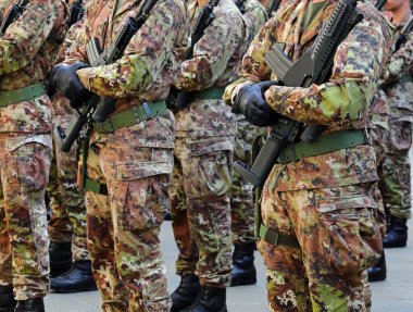 Yeşil ve kahverengi kamuflaj üniformalı askerler eldiven ve saldırı tüfekleri ellerinde.