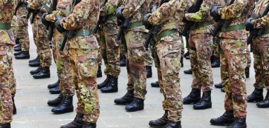 Yeşil ve kahverengi kamuflaj üniformalı askerler. Kışlada siyah asker botları ve saldırı tüfekleri var.