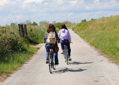 Genç anne ve kız yazın nehir kıyısının yanındaki sırt çantasıyla bisiklet yolunda pedal çeviriyorlar.