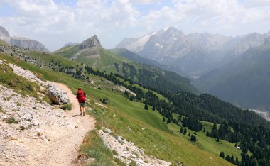 Omuzlarında kırmızı sırt çantasıyla dağ yolunda yürüyen ve arka planda 3300 metre irtifada Marmolada buzulu olan genç yürüyüşçü.