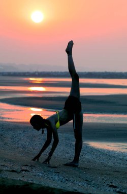 very flexible and athletic girl in backlight doing gymnastics on the seashore in summer raising her leg vertically with a yellow bra clipart