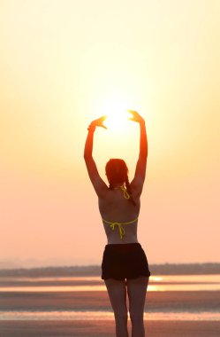 Arkasından kolları havada, kumsalda elleri ile güneşi yakalamaya çalışan bir kız.