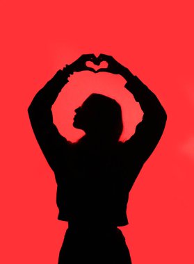 suggestive black silhouette of the girl in profile who with her hands forms a heart on the very red background clipart