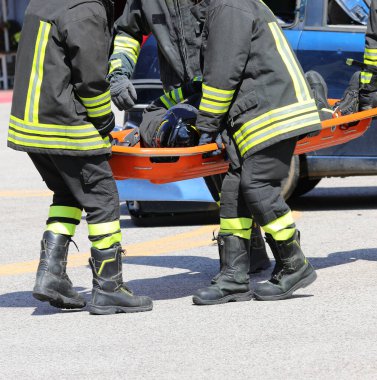 Üniformalı itfaiyeciler acil durum sahnesinde bir kişiyi turuncu sedyeye kaldırarak kurtarma operasyonu gerçekleştiriyorlar.