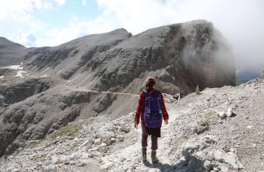 young woman walks with backpack on shoulders in rugged path of stony mountain in European Alps in summer clipart