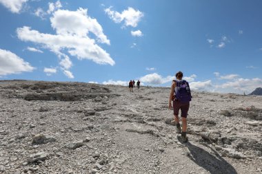 young female hiker with purple backpack on shoulders and cap walking on stony path in european alps in northern italy in summer clipart