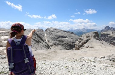 young woman hiker with a backpack on her shoulders points at mountain peaks clipart