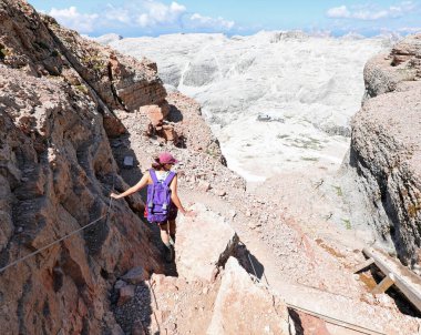 Genç bir kadın dik bir dağ yolunda, sırtında bir sırt çantasıyla, güvenlik için ferrata geçidinin çelik kablosuna sıkıca tutunuyor.