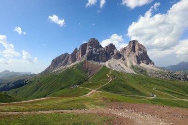 Panoramic view of the Italian Dolomites with the Sasso Lungo mountain group and many trails in the green meadow below during summer clipart