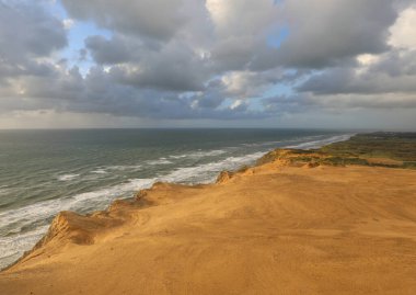 Kumlu sahil şeridi giderek denizin gücüyle aşınıyor, bulutlu bir gökyüzünün altında, iç çeken insanlar olmadan.