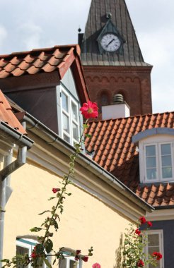 Kırmızı çiçek ve İskandinav tarzı evlerin eğimli çatıları. Çan kulesinde analog bir saat var.