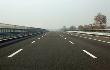 Her iki yönde üç şeritli, hafif sisli bir düzlükten geçen az trafiği olan düz bir karayolu.
