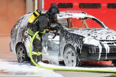 Üniformalı itfaiyeci ve kendine yeten solunum cihazıyla araba kazasından sonra çıkan yangını söndürmek için beyaz köpük püskürtüyor.