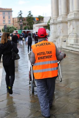 Avrupa seyahati sırasında yolcuların bavullarını taşımasına yardım etmek için floresan turuncu yelekli kapıcı.