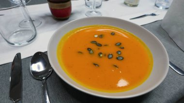 Bir kase kremalı portakallı balkabağı çorbası. Üstünde çıtır çıtır çekirdekler var. Masada güzelce servis ediliyor. Parlak renk ve kadifemsi doku. .
