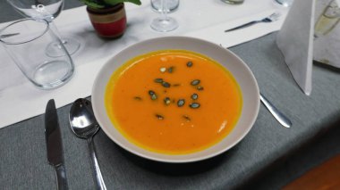 Bir kase kremalı portakallı balkabağı çorbası. Üstünde çıtır çıtır çekirdekler var. Masada güzelce servis ediliyor. Parlak renk ve kadifemsi doku. .