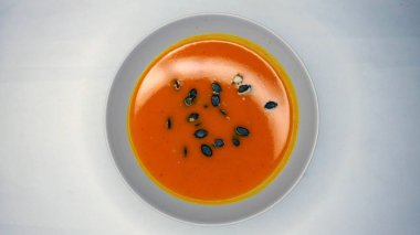 Bir kase kremalı portakallı balkabağı çorbası. Üstünde çıtır çıtır çekirdekler var. Masada güzelce servis ediliyor. Parlak renk ve kadifemsi doku. .