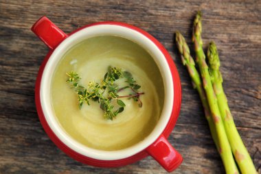 Kırmızı bir kasede kuşkonmaz çorbası, yanında taze sebzeler ve ahşap bir arka plan.